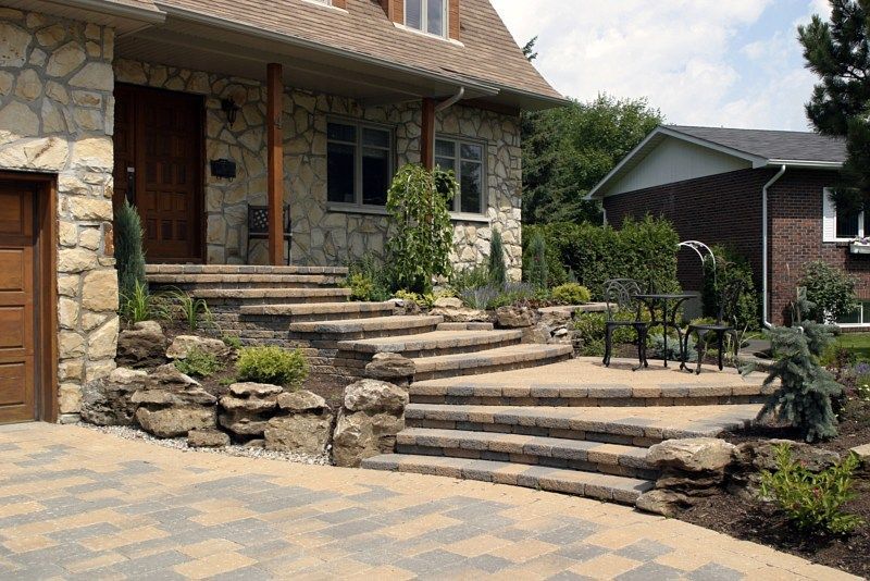 A stone house with an outdoor patio featuring tiered stone steps, a paved driveway, and landscaping with large rocks.
