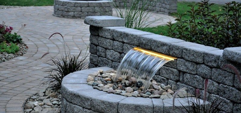 A stone retaining wall with a lighted waterfall flowing into a circular stone basin in a landscaped patio.