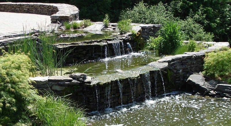 A tiered stone water feature with small waterfalls cascading into a garden pond surrounded by lush green plants.