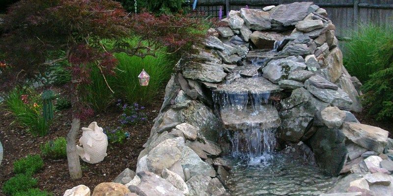A tiered stone waterfall feature flowing into a small pond, situated in a garden with a Japanese maple and greenery.