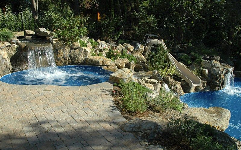 A landscaped backyard pool area with waterfalls, rock features, and a water slide connecting a spa to a larger pool.