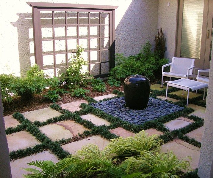 A modern courtyard features square stone pavers separated by grass, a black stone fountain, and contemporary patio chairs.