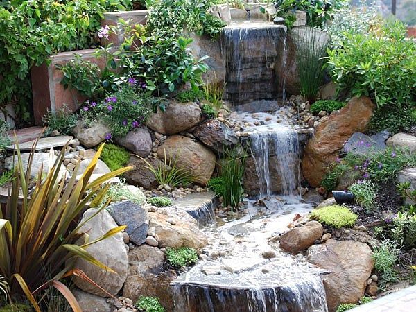 A tiered stone waterfall flows into a lush garden with rocks, green foliage, and flowering plants.
