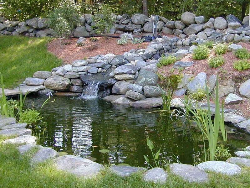 A small backyard pond with a cascading stone waterfall, surrounded by green grass, mulch, and natural landscaping rocks.