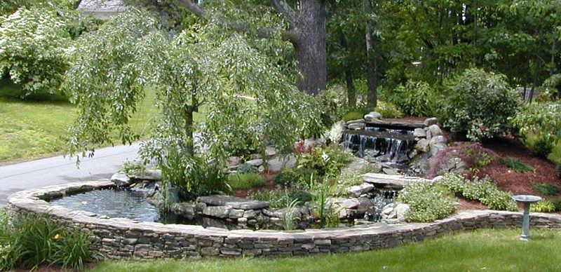 A stone-walled koi pond with a small waterfall, surrounded by lush green plants, trees, and landscaping.