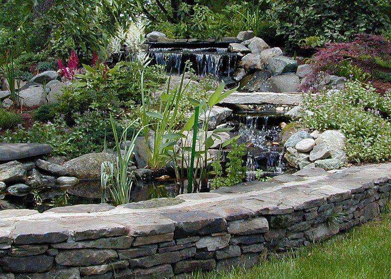 A tiered stone garden pond with a cascading waterfall, lush greenery, and a low stacked-stone retaining wall.