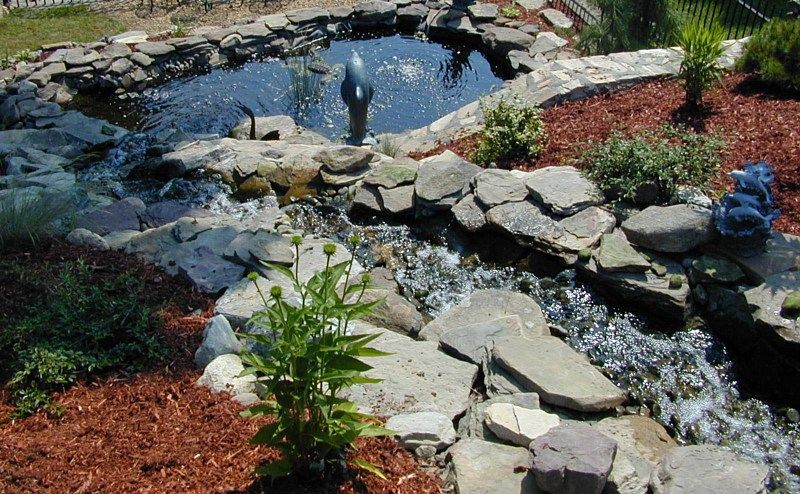 A tiered backyard water feature with a stone-lined stream flowing into a small pond surrounded by mulch and greenery.