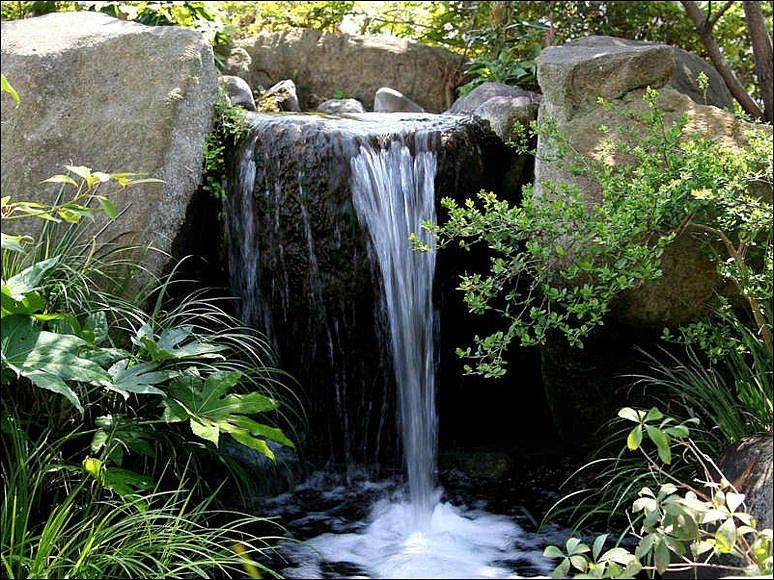 A small, tiered garden waterfall framed by large gray rocks and lush green foliage.