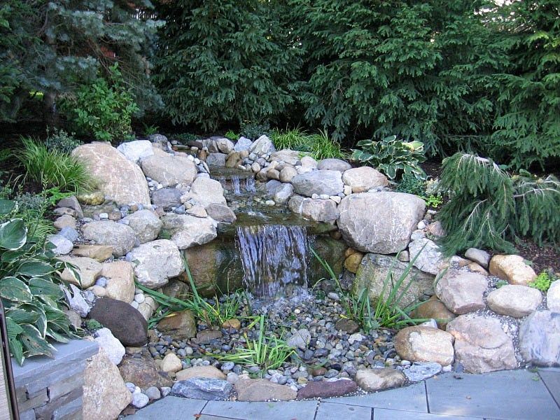 A small backyard waterfall flows over natural stone into a gravel-filled basin, surrounded by lush green foliage.