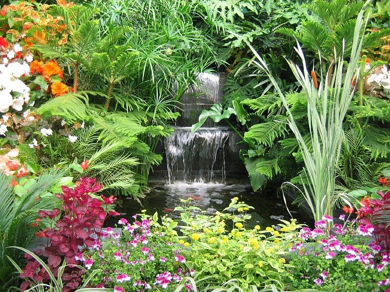 A tiered garden waterfall flowing into a dark pond, surrounded by lush green foliage and colorful flowering plants.