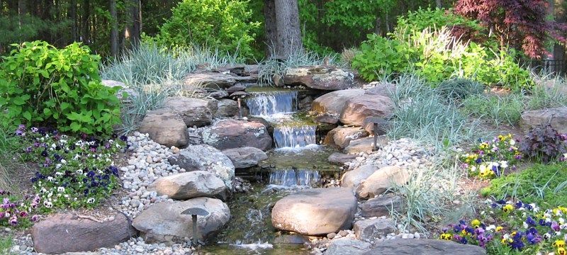 A small, tiered stone waterfall flows into a shallow stream surrounded by various green plants and colorful flowers.