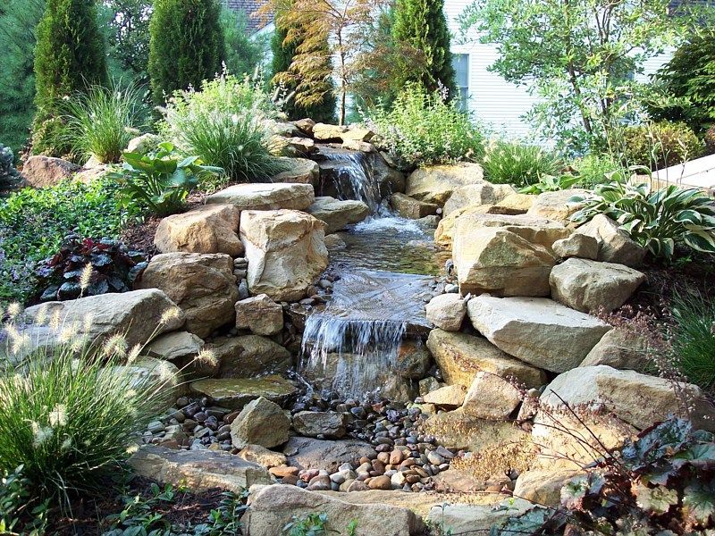 A tiered garden waterfall cascades over large, stacked natural rocks surrounded by lush greenery and ornamental grasses.
