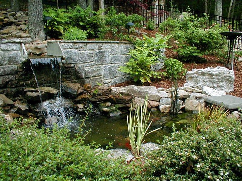 A stone waterfall flows into a backyard pond, surrounded by lush green plants and landscaping.
