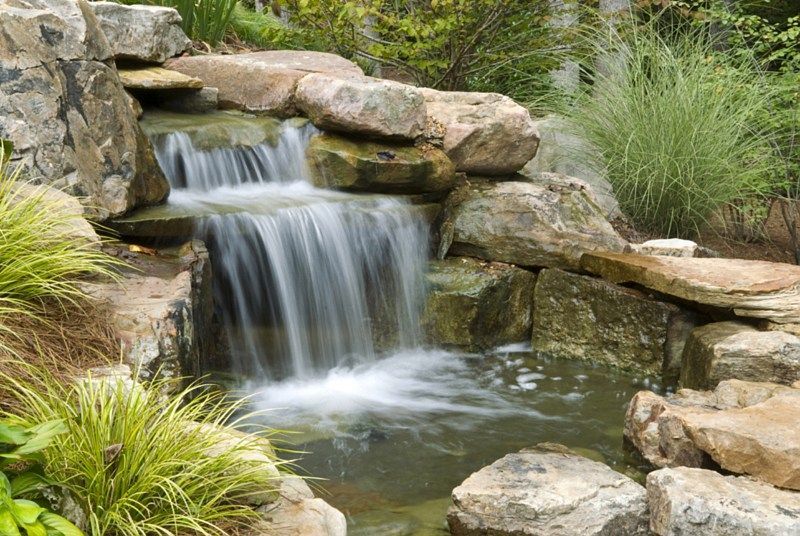 A tiered rock garden waterfall flowing into a small pond, surrounded by lush green foliage and stone landscaping.