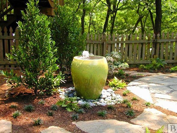 A lime green ceramic fountain vase sits on a bed of river rocks in a garden, near a flagstone path and a wooden fence.