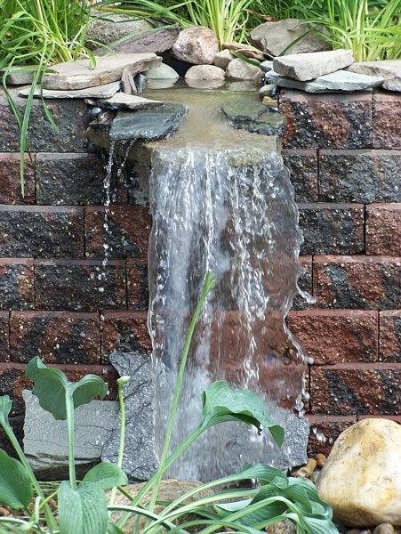 A small stone waterfall flowing over a brick retaining wall into a garden, framed by green plants and smooth rocks.