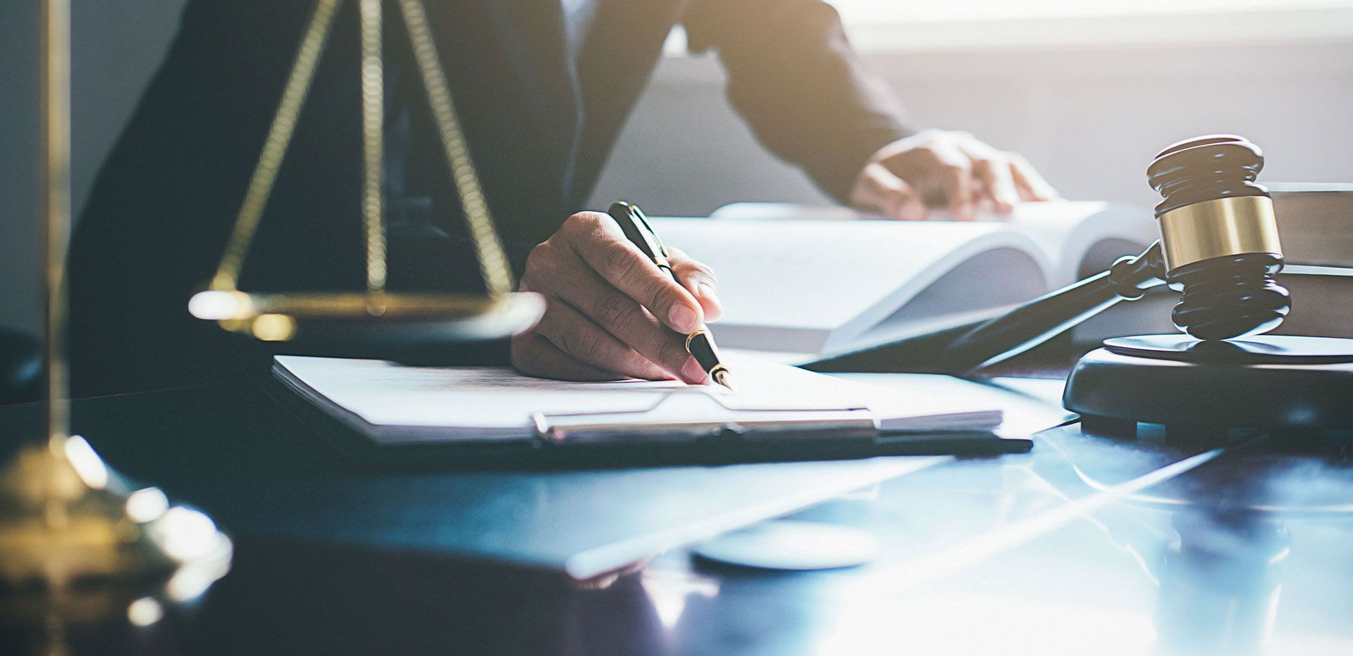 A person writing on a document with a pen, next to a gavel, scales of justice, and open book on a desk.