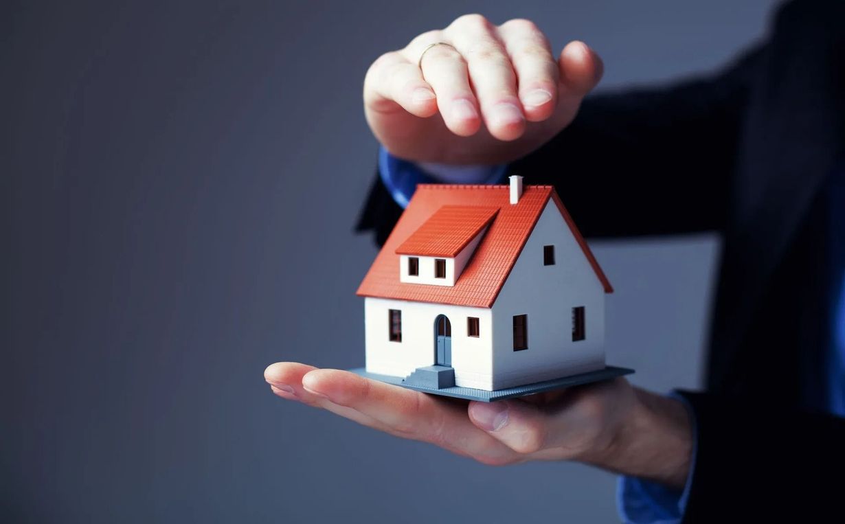 Hand holding a model house, other hand above in a protective gesture, against a gray background.