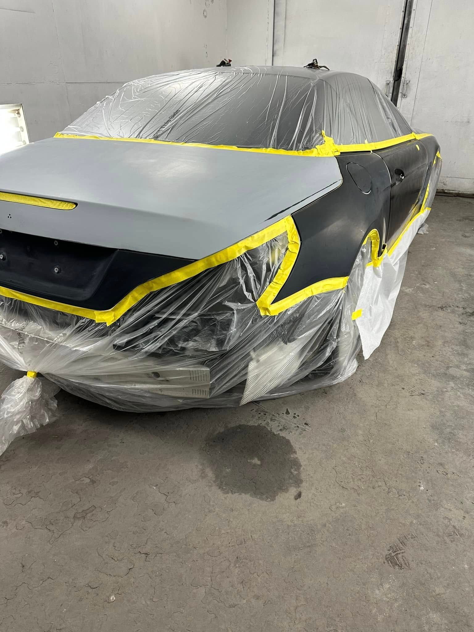 A black car is sitting in a garage covered in plastic wrap.