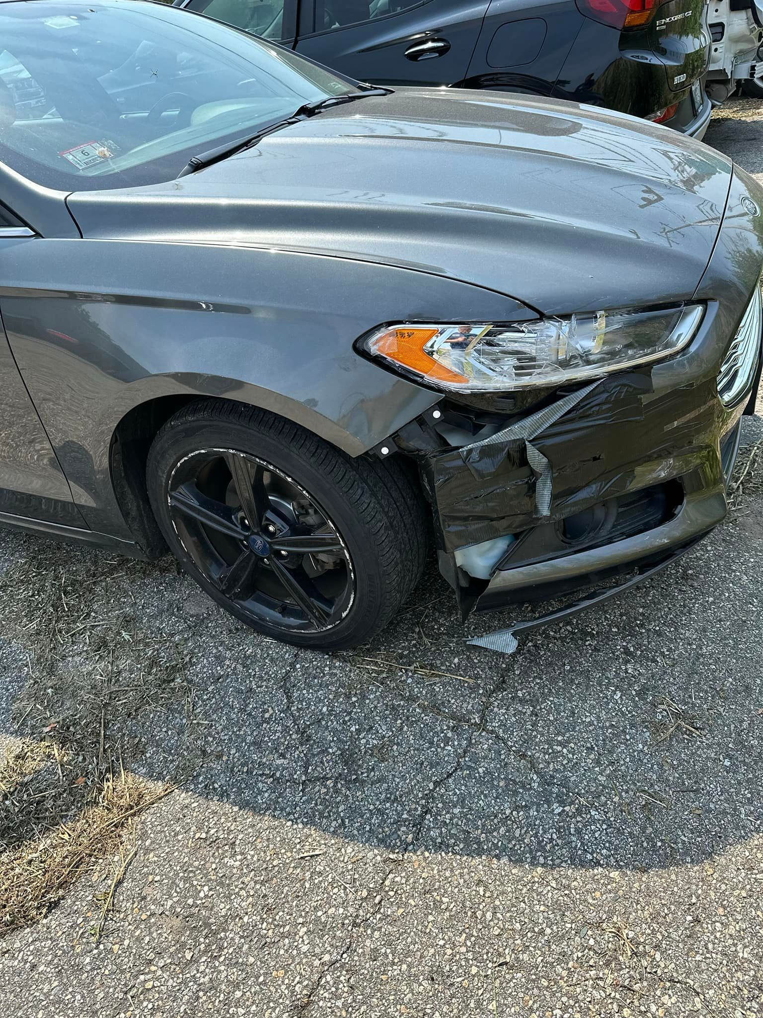 A gray car with a damaged front bumper is parked on the side of the road.