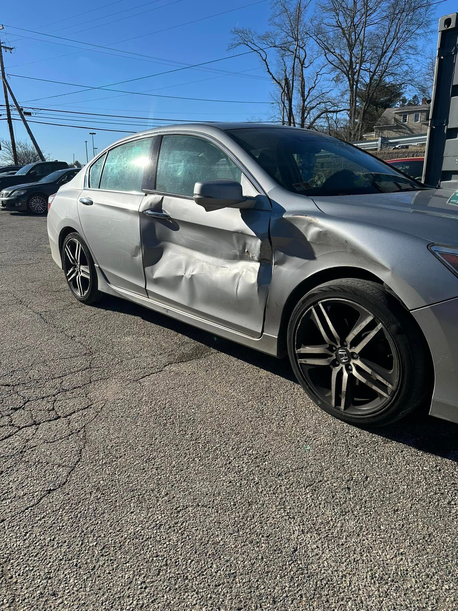 A silver car with a damaged side is parked in a gravel lot.