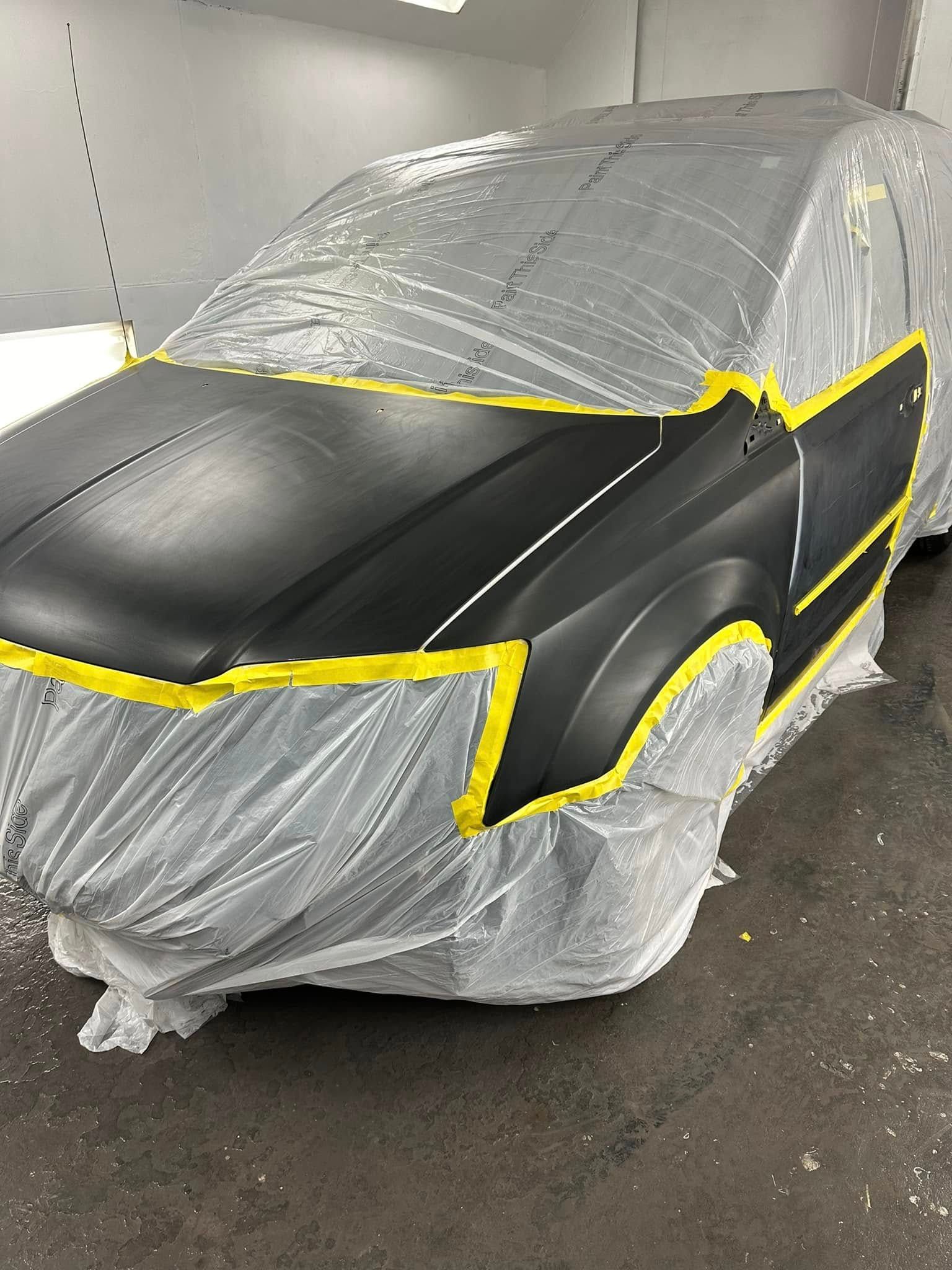 A black car is covered in plastic and tape in a garage.