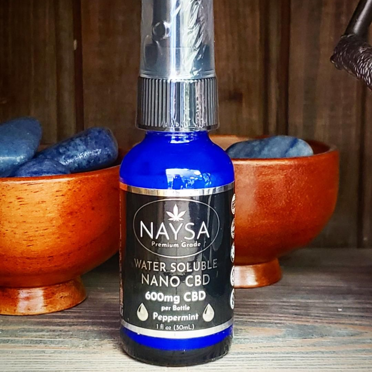Blue Naysa water-soluble nano CBD bottle with silver cap on a wooden surface beside bowls and stones.