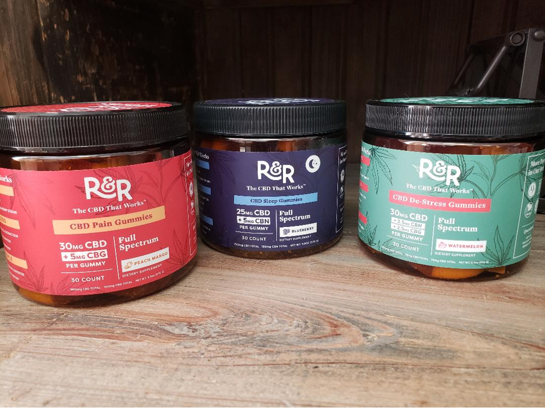 Three jars of R&R CBD topical creams in red, blue, and green with black lids, displayed on a wooden shelf.