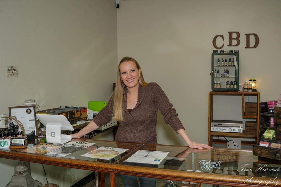 Woman smiling behind a CBD shop counter, with products displayed and 