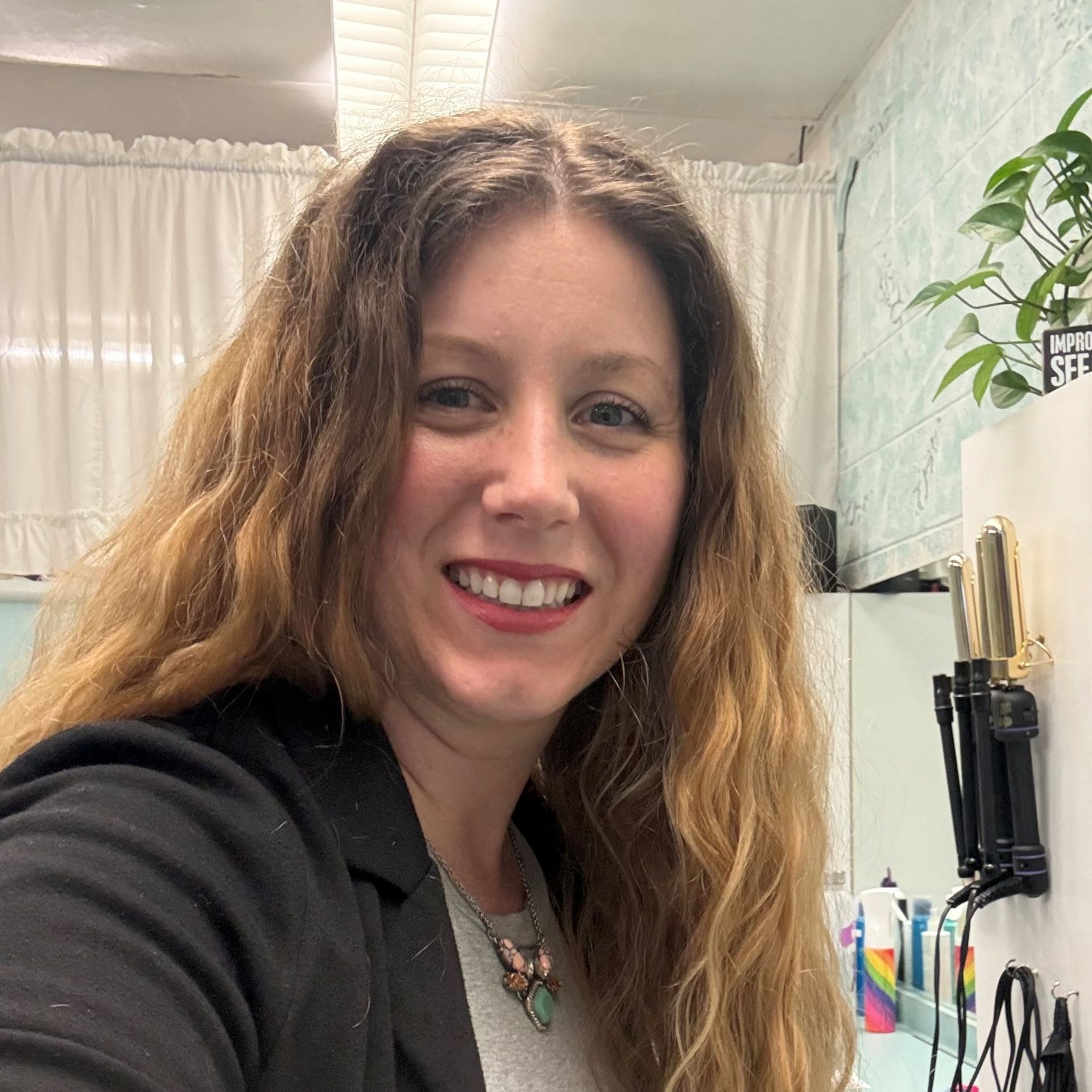 A smiling person with wavy, light-brown hair, wearing a black blazer, poses in a salon with styling tools in the background.