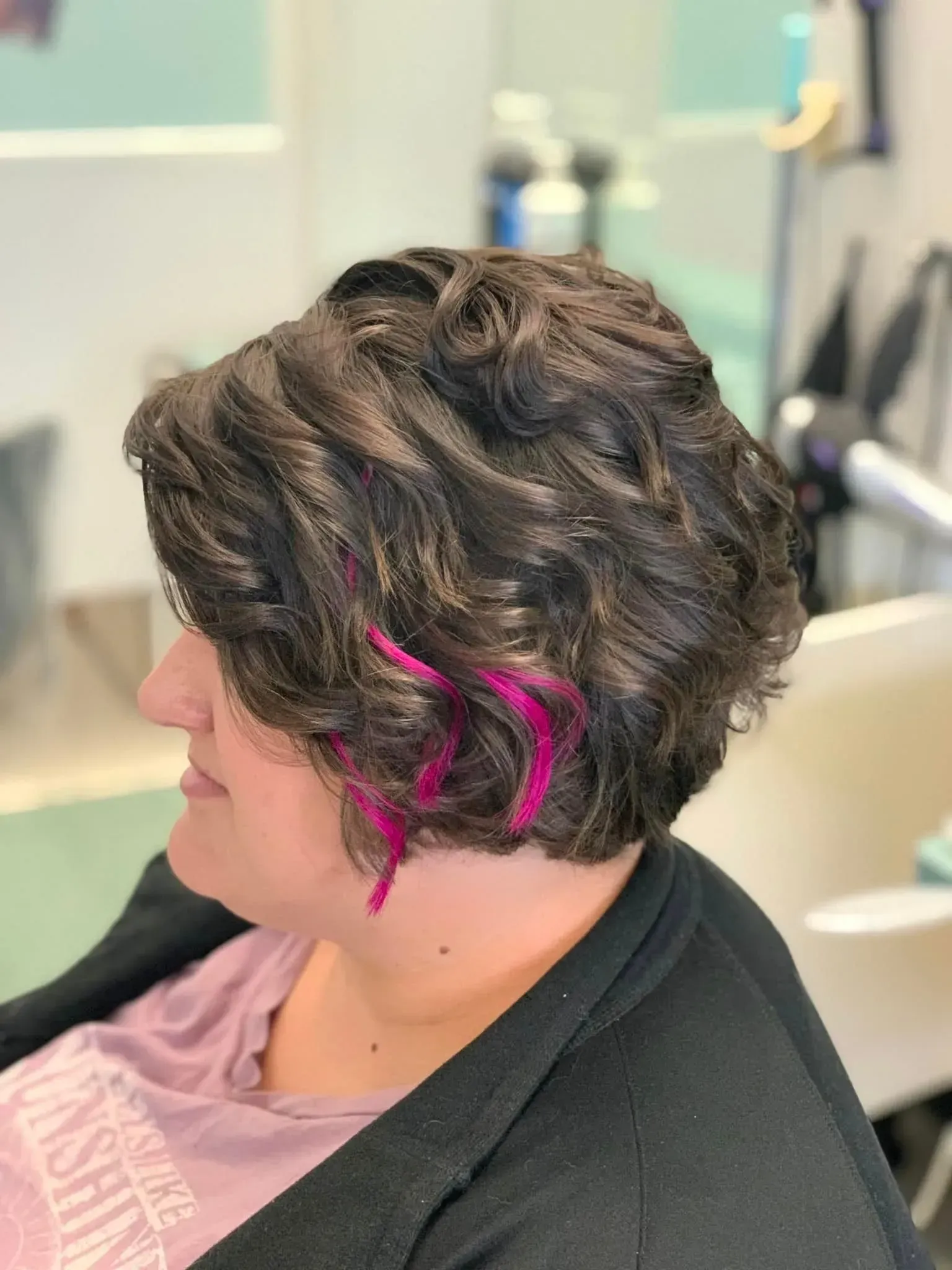 Woman with dark, wavy hair, pink highlights, in salon setting.