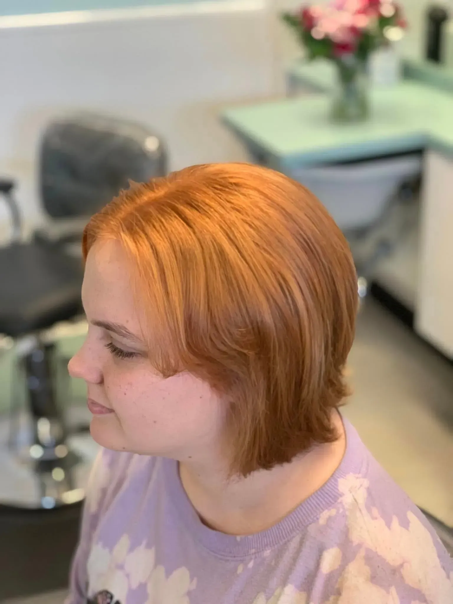 Woman with short, reddish-orange hair at a salon, wearing a lavender shirt, looking left.