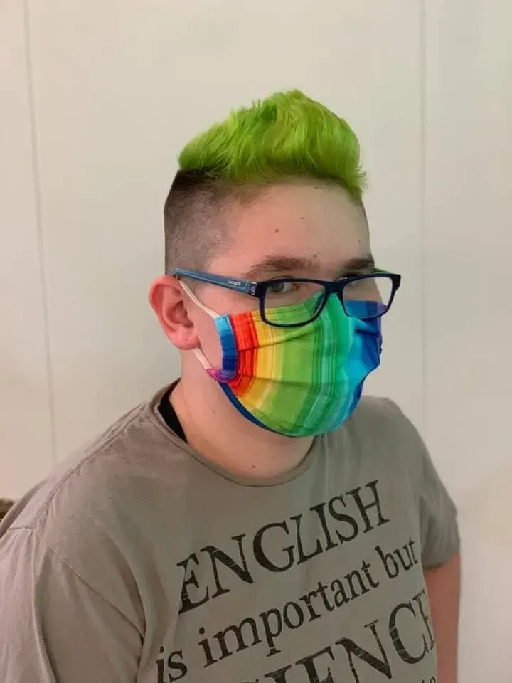 Person with green hair and rainbow mask, wearing glasses, in front of a white wall.