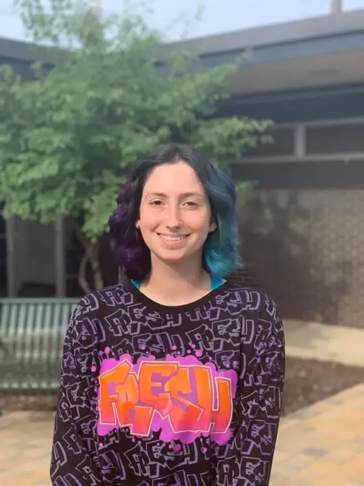 Woman with blue and purple hair smiles, wearing a black sweatshirt with colorful graffiti art; outdoors, near a building.