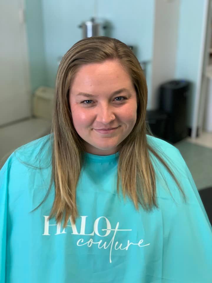 Woman with long blonde hair wearing a teal Halo Couture salon cape; smiling indoors.