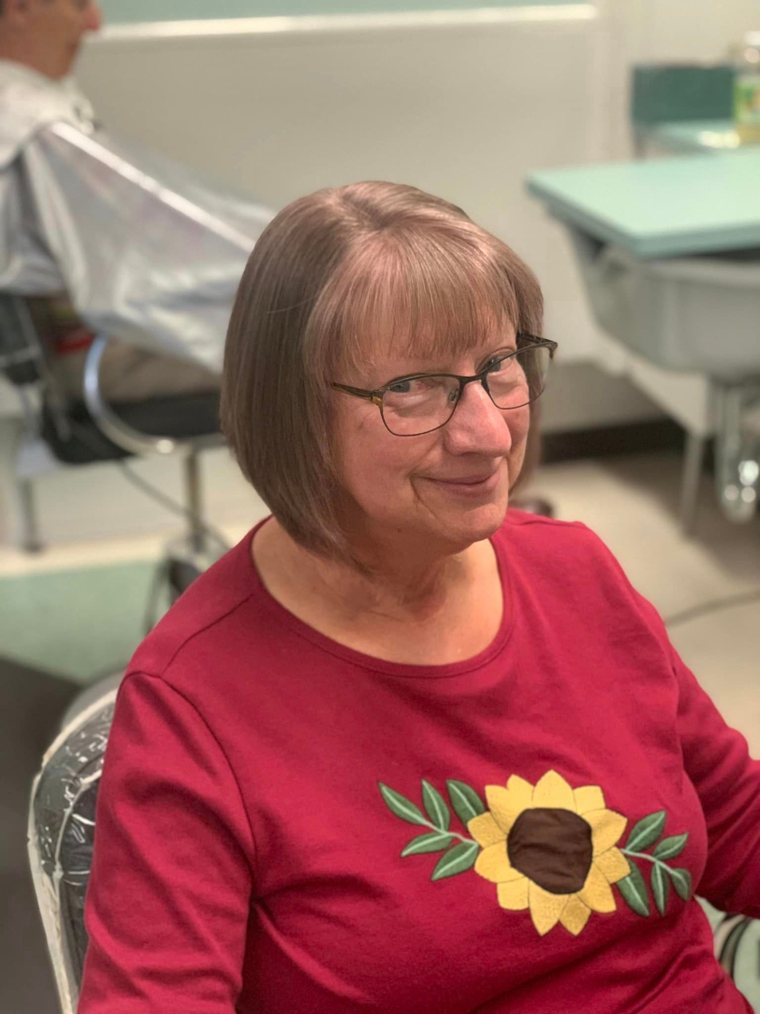 Woman with a new haircut, wearing a red shirt with a sunflower. Sitting in a salon chair.