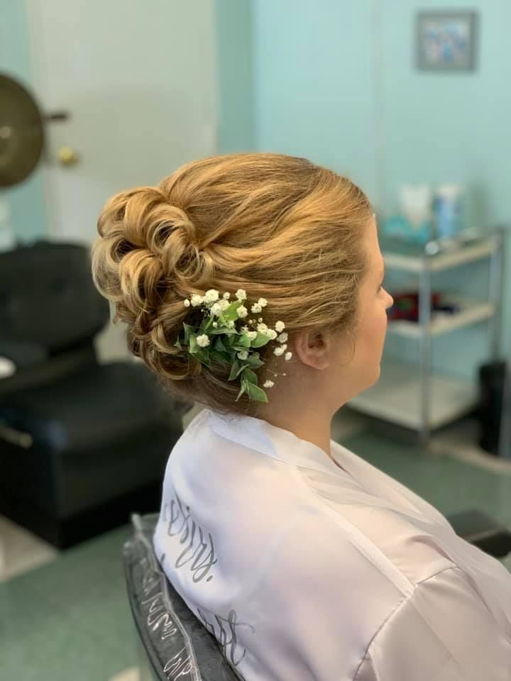 Woman with blonde updo decorated with flowers, wearing a white robe, in a salon.