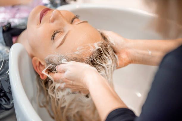 Woman getting her hair washed at a salon, head in a sink, eyes closed, blonde hair covered in shampoo.