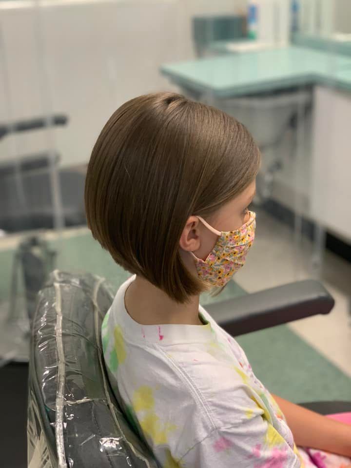 Girl with a short, layered bob haircut, wearing a floral face mask, sits in a salon chair.