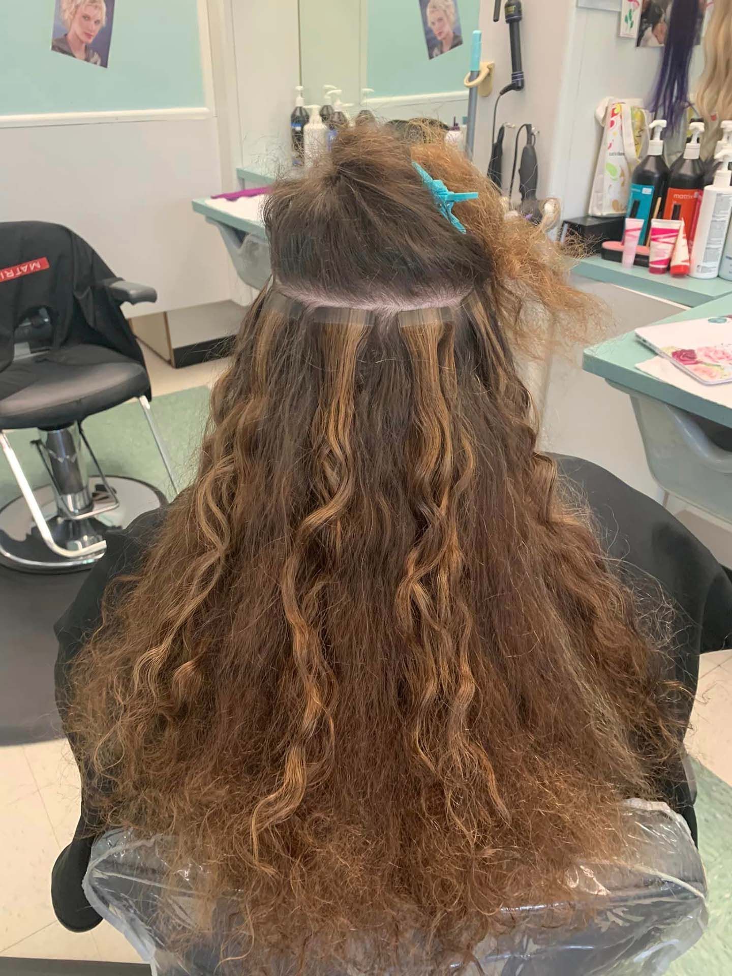 A person in a salon chair with hair extensions partially installed; brown wavy hair, a blue clip, salon setting.