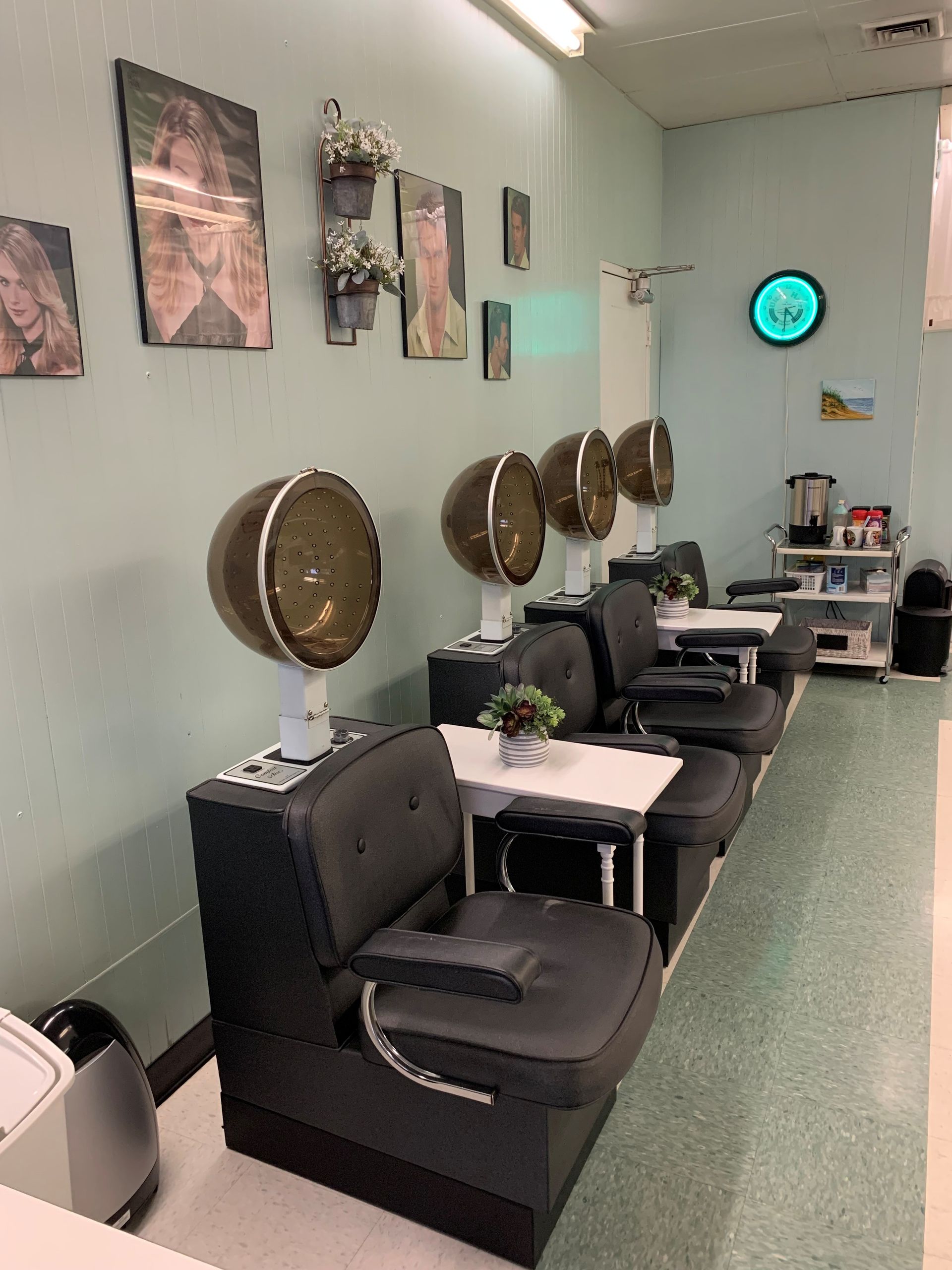 A row of four salon chairs with professional hooded hair dryers, positioned along a light green wall with framed art.