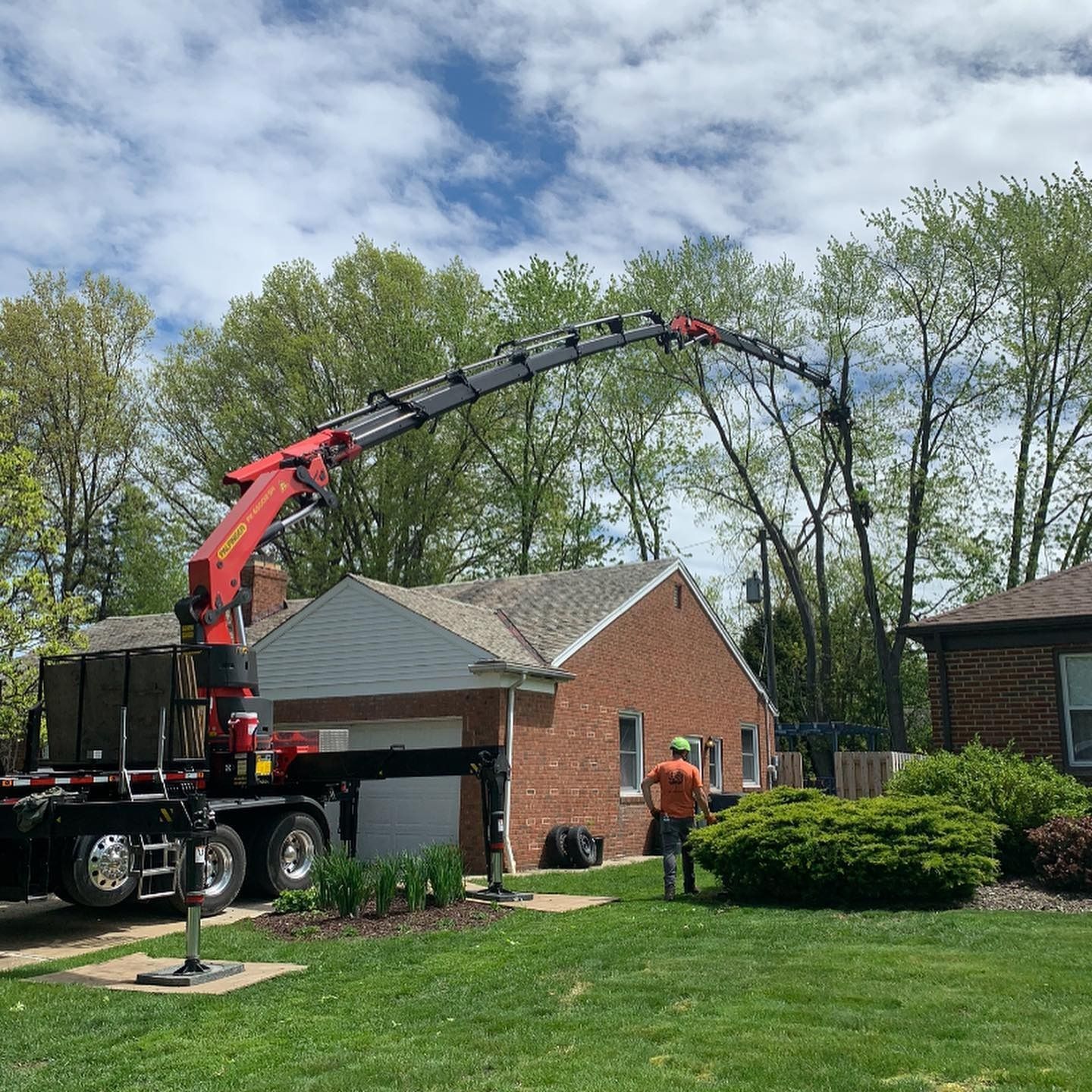 A red boom crane parked on a lawn extends its arm toward tree branches near a brick house while a worker stands nearby.