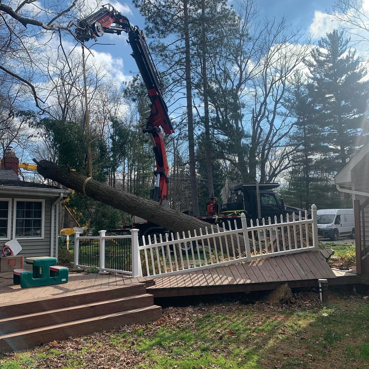 A crane removes a fallen tree trunk from above a deck and a white picket fence at a house.