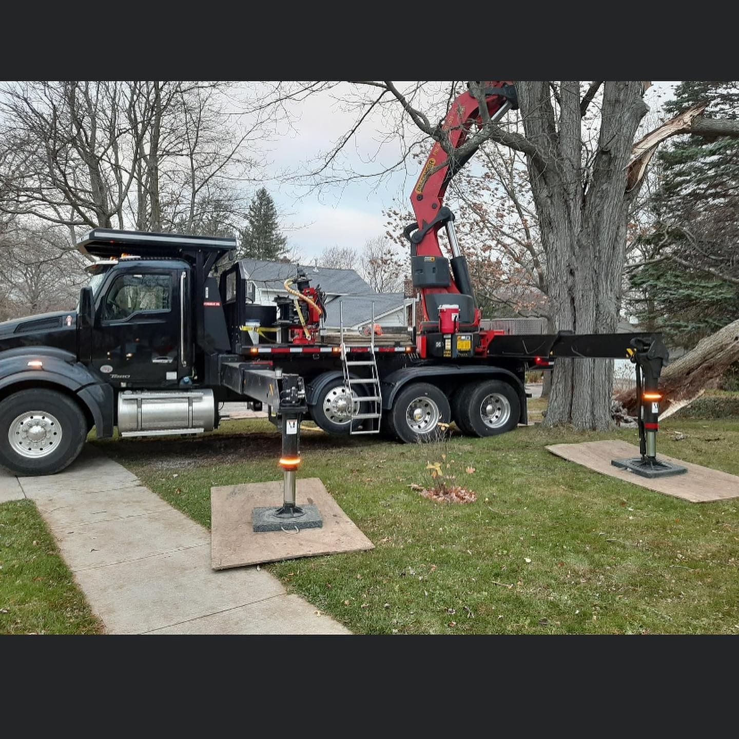A black crane truck with extended outriggers parked on a grassy lawn next to a tree.