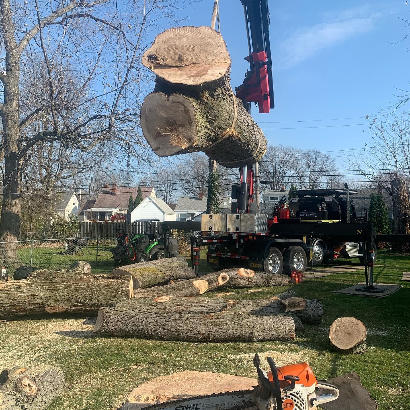 A crane lifts a large, cut tree trunk over a residential yard filled with additional logs and a chainsaw.