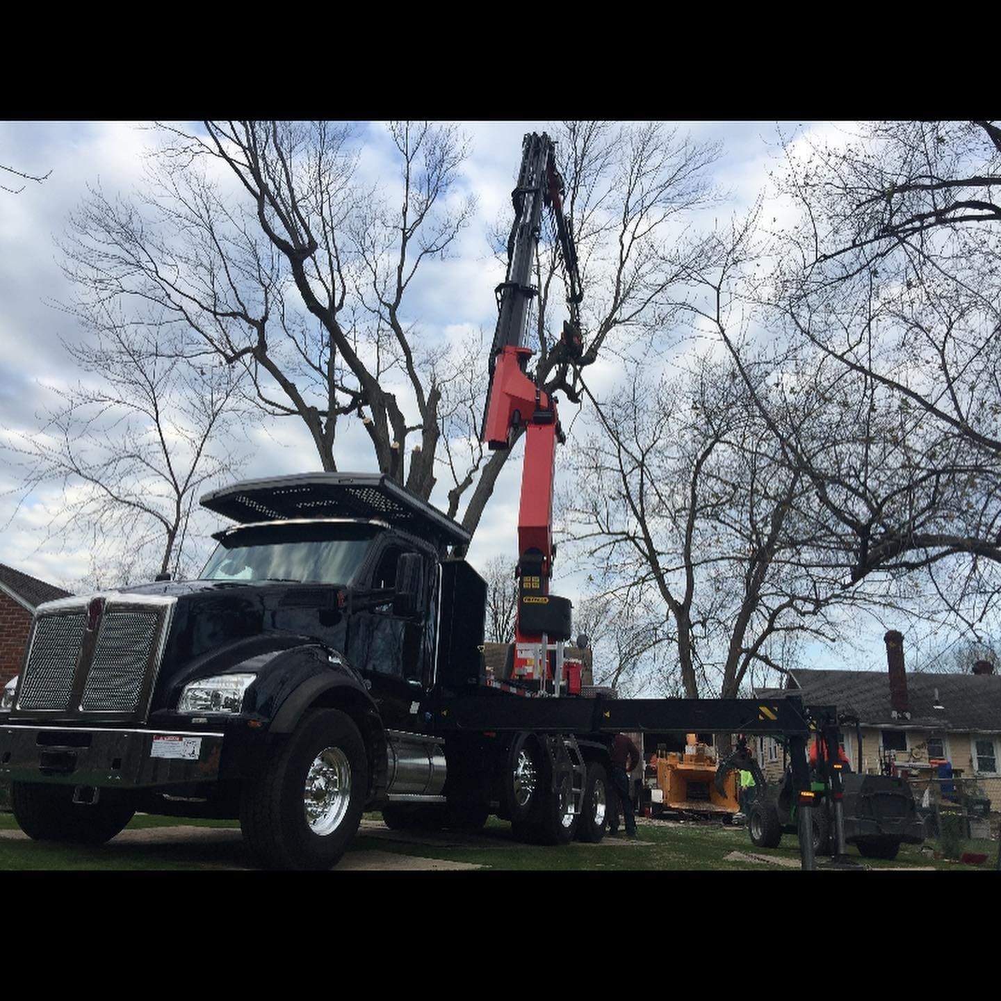 A black Kenworth truck with a red grapple crane arm raised high, working on tree maintenance in a residential yard.