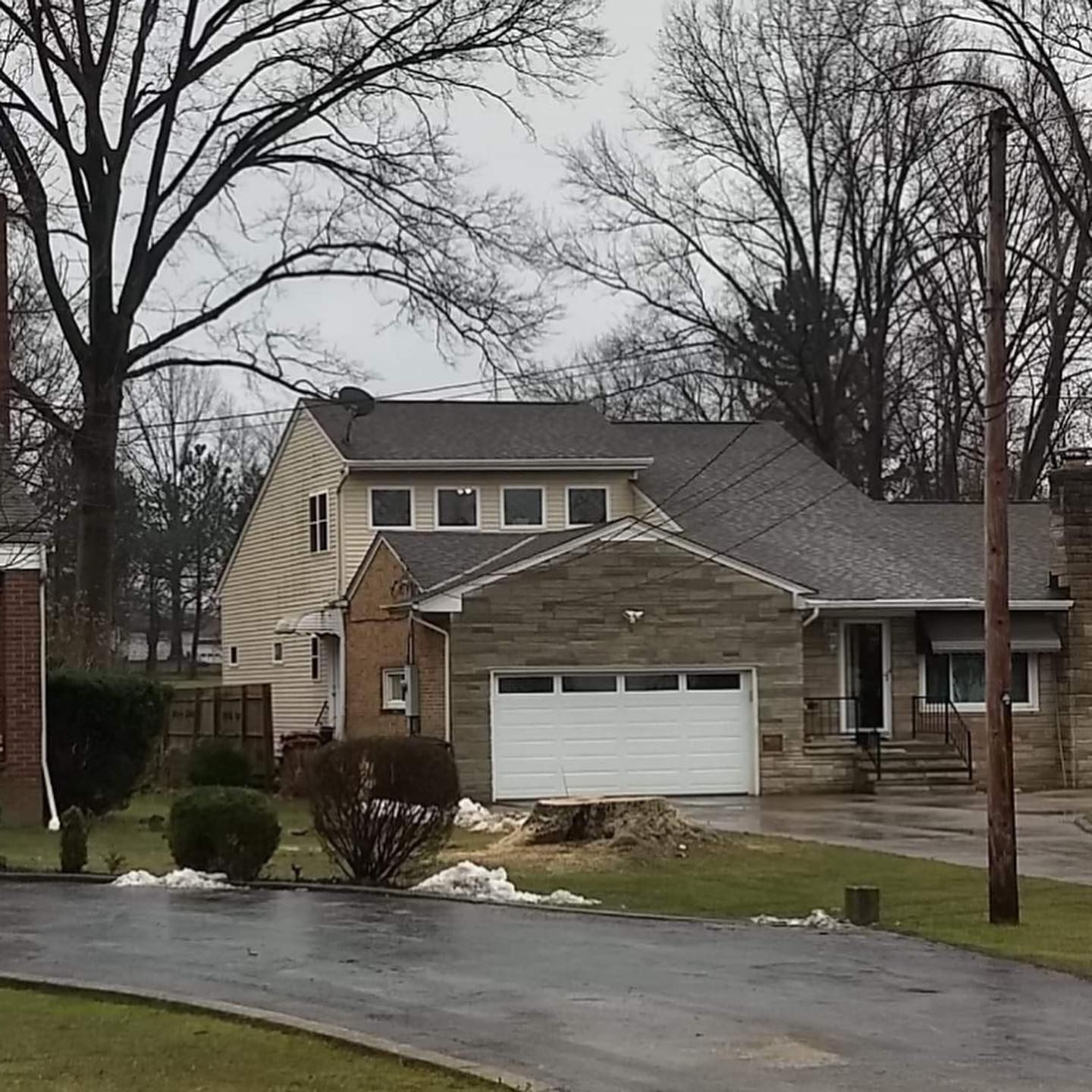 A two-story suburban house with a stone-front garage, tan siding, and a paved driveway on an overcast day.