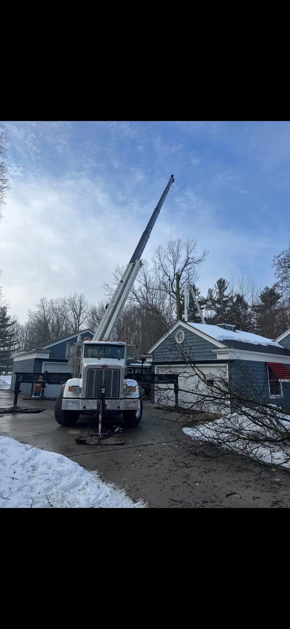 A white crane with an extended boom stands in a snowy residential driveway next to a house with downed tree branches.