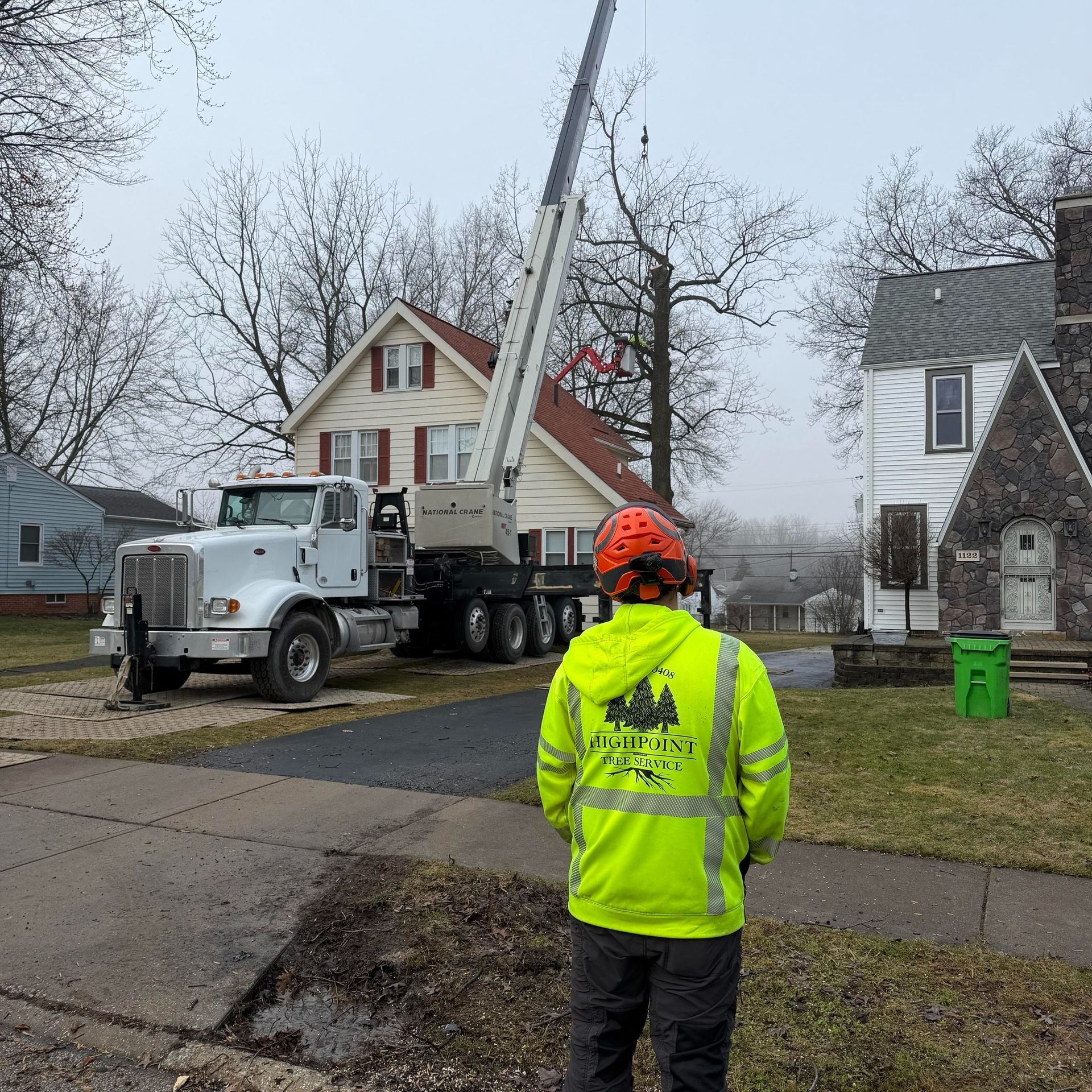 A worker in a bright yellow safety jacket observes a large crane lifting tree limbs near residential houses.