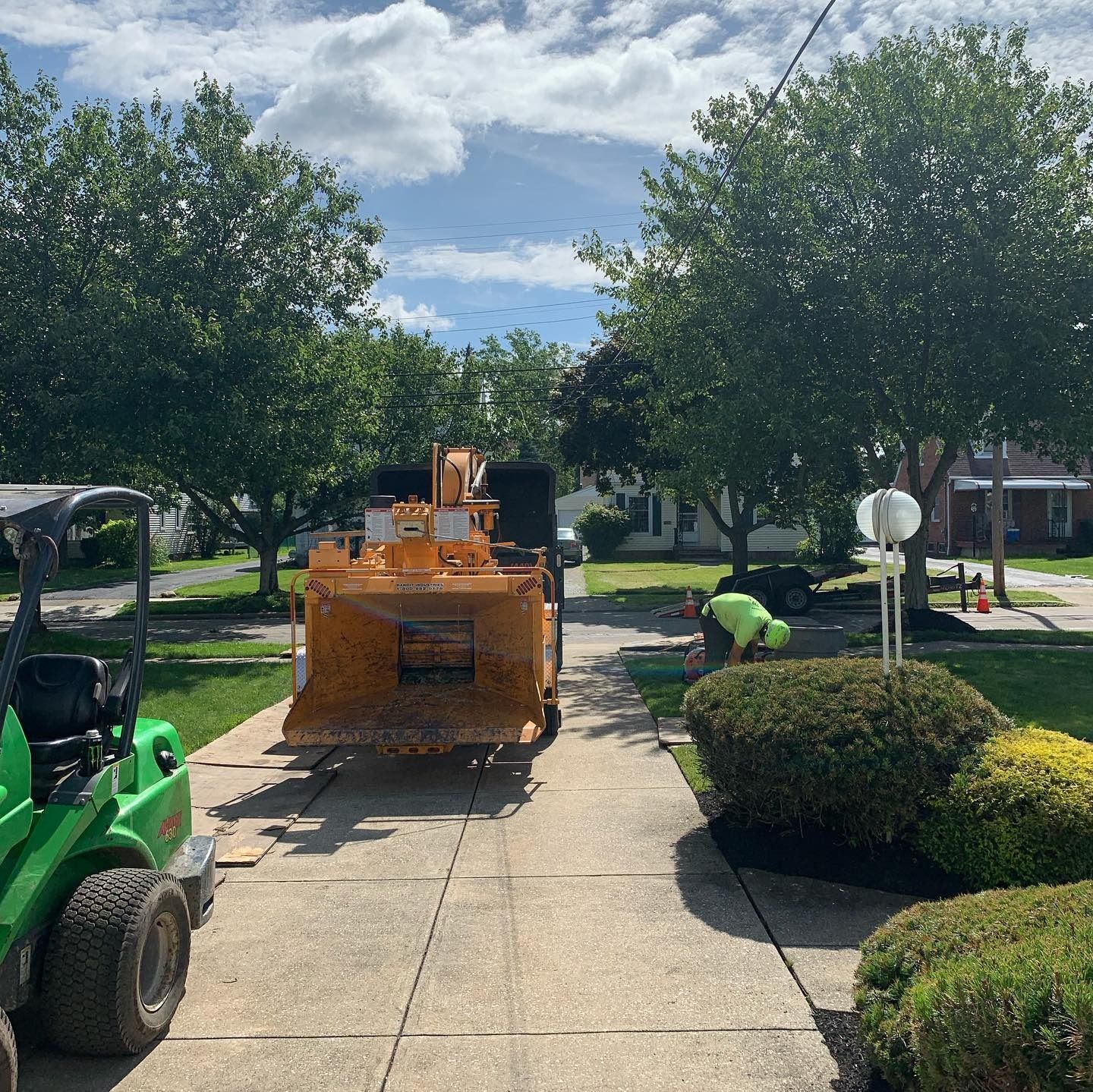 A green skid steer and a large yellow wood chipper parked on a residential driveway on a sunny day.